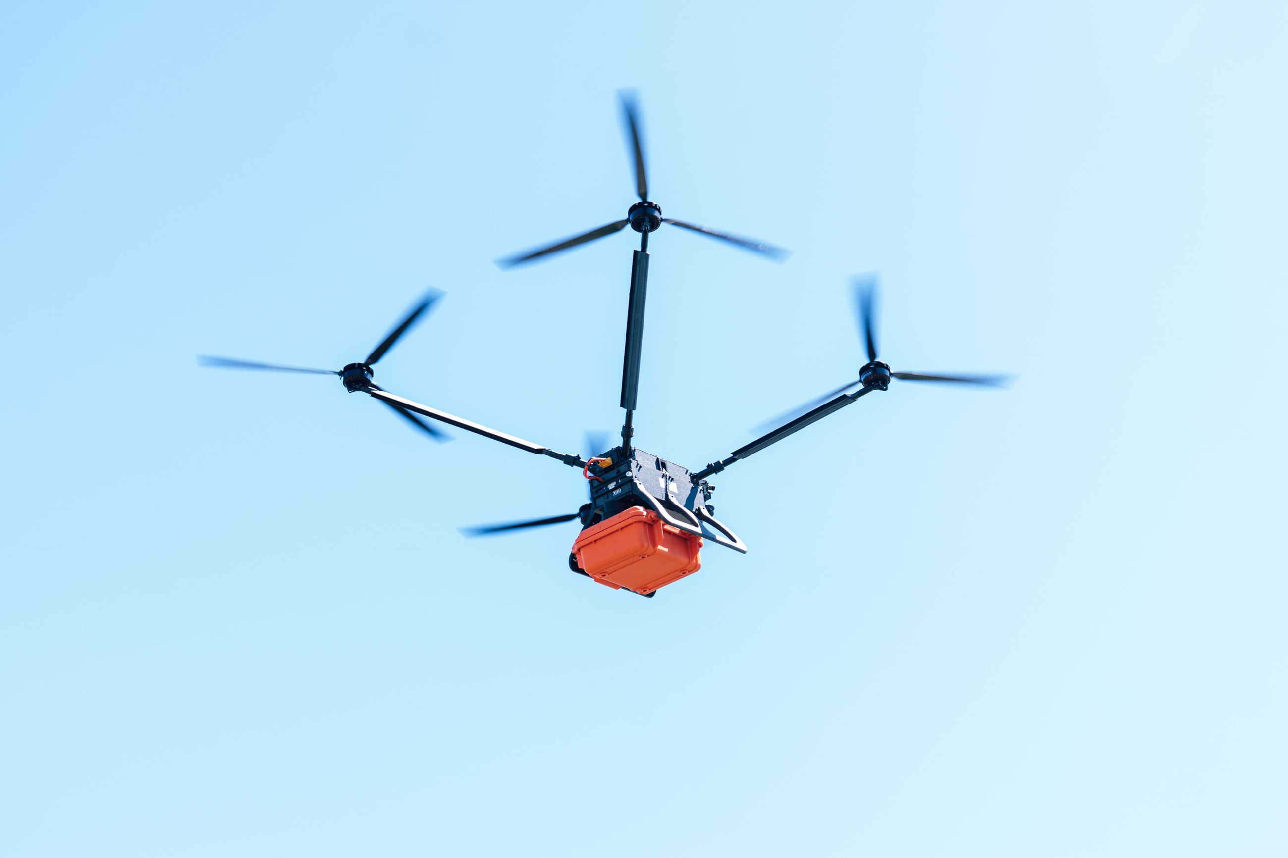 Black quadcopter drone with an orange cargo box flying against a clear blue sky.