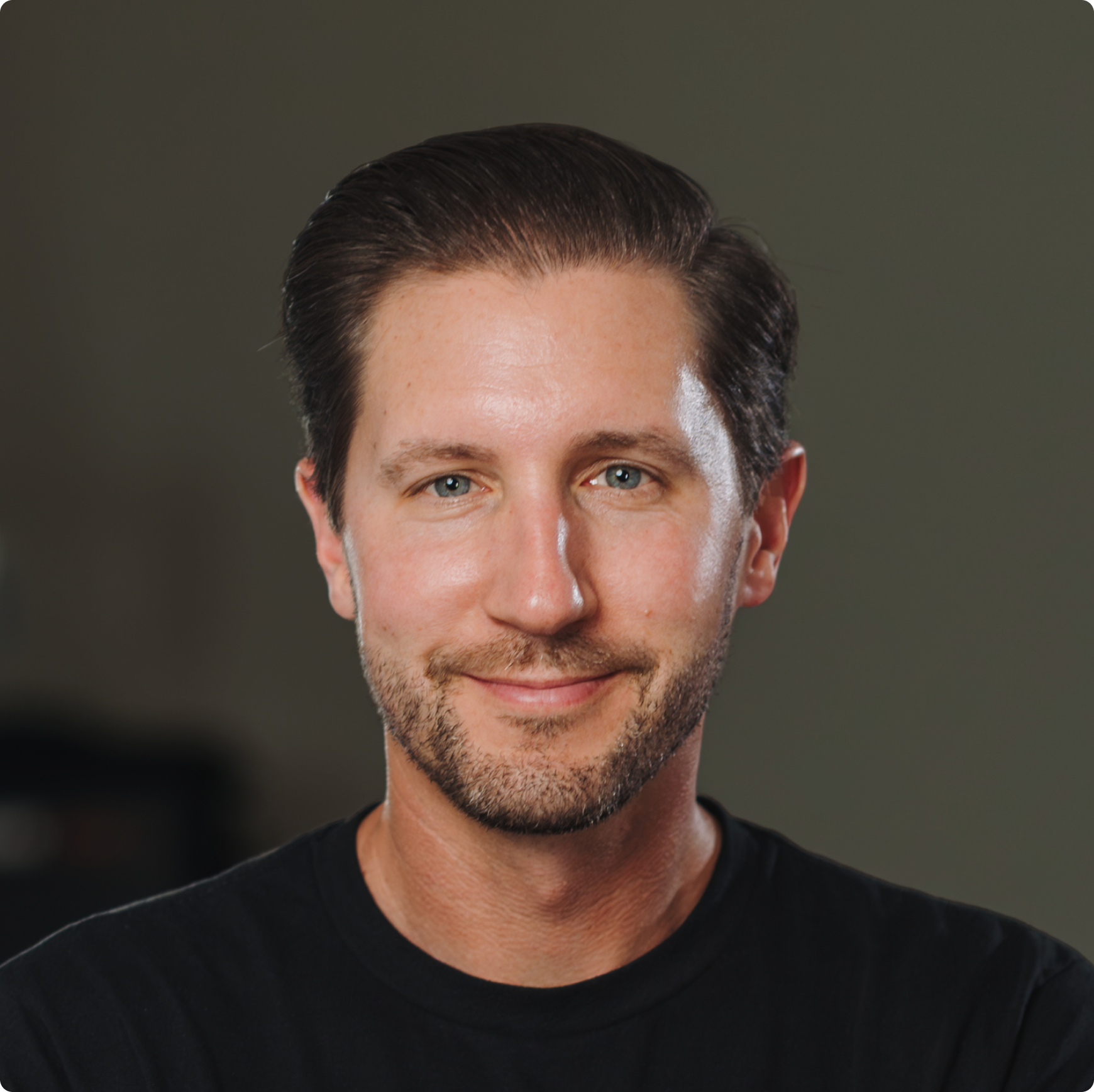 Smiling man with short brown hair, beard, and blue eyes wearing a black shirt against a plain background.
