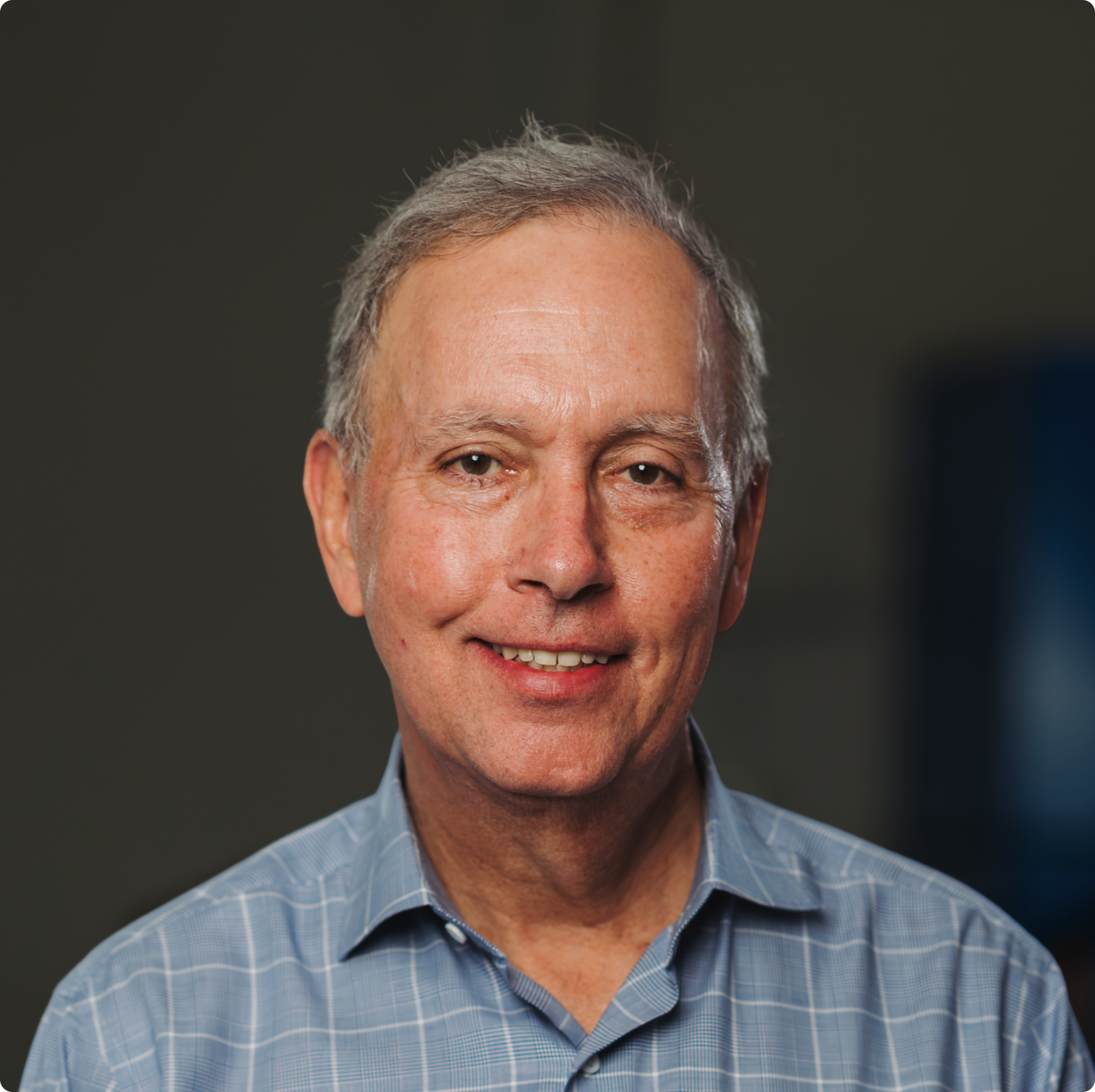 Smiling middle-aged man with gray hair wearing a light blue checkered shirt against a dark background.